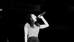 A black-and-white image. Against a black background, a young woman in side profile holds a microphone high and belts into it.