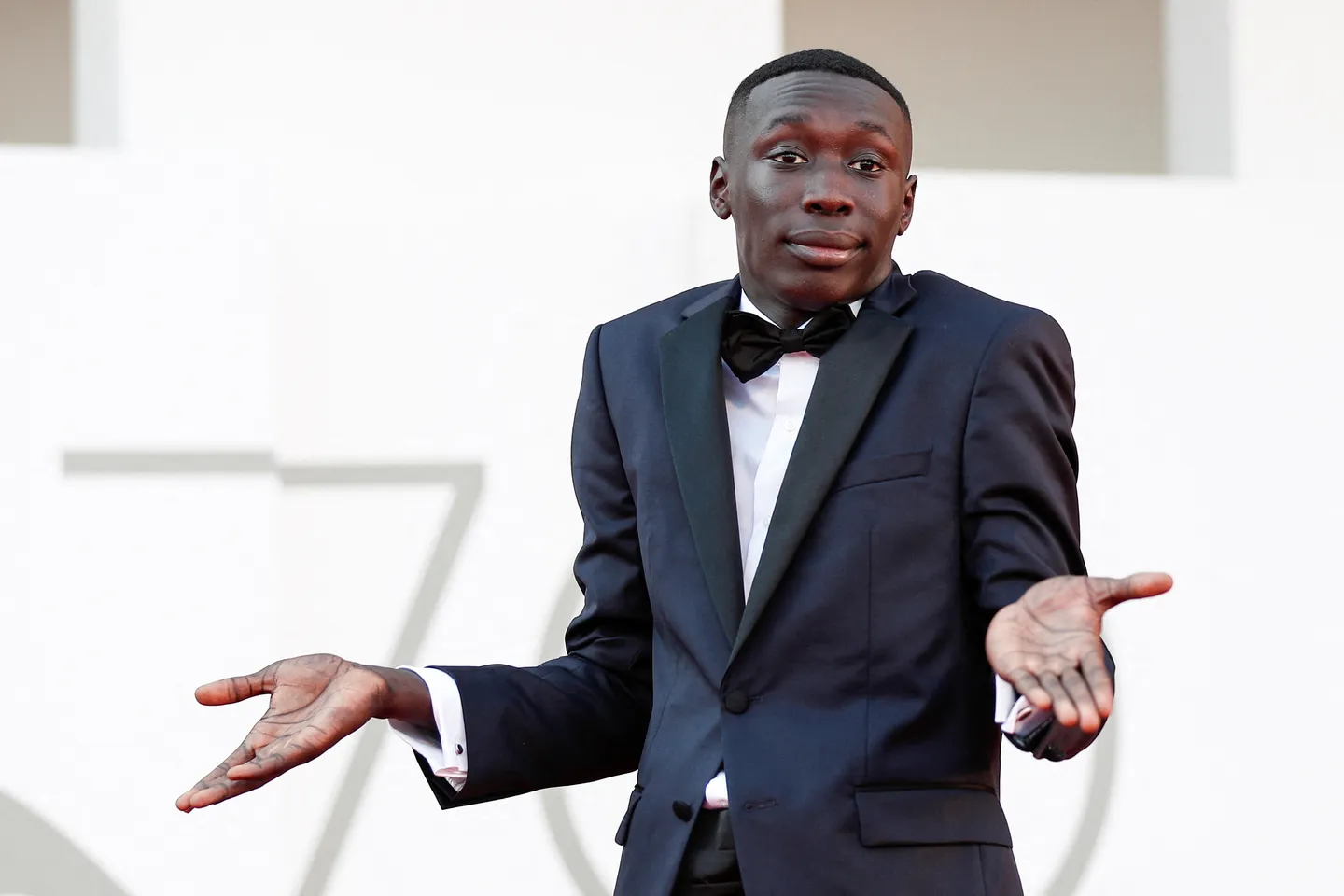Khaby Lame wearing a black tuxedo, performing his signature two-hand gesture with a neutral expression.