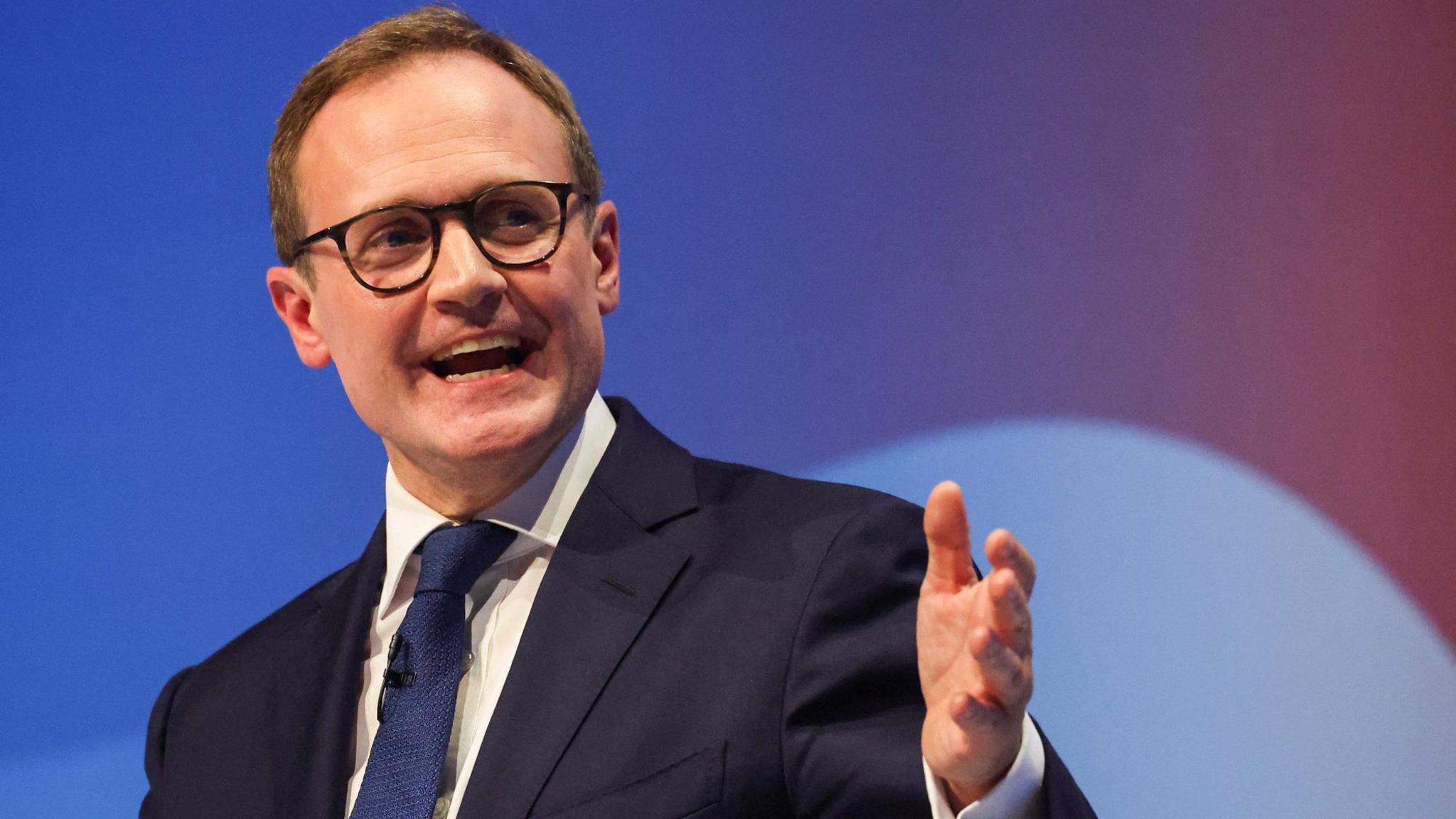 Tom smiling warmly while giving a speech on stage in front of an audience.