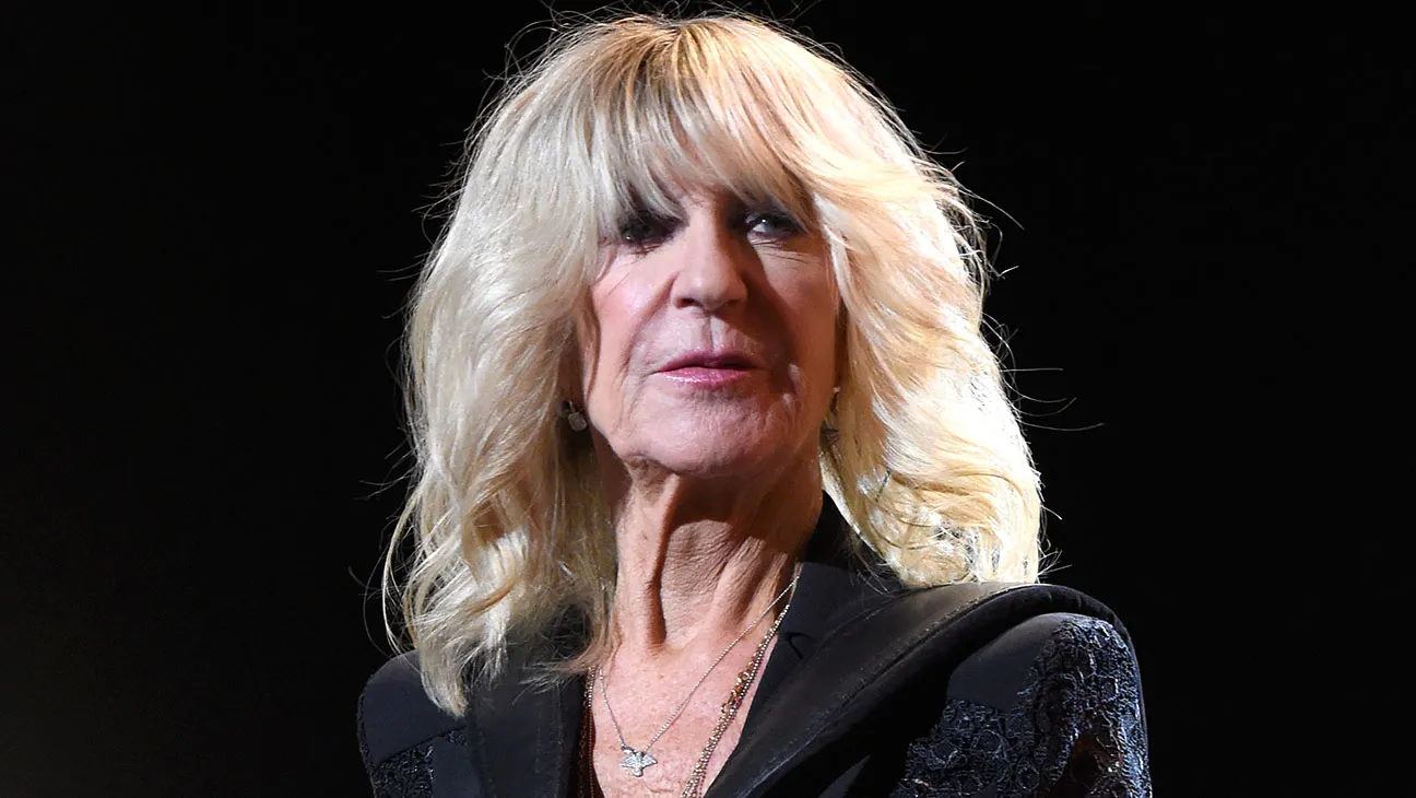 Christine McVie performing on stage, playing the keyboard and singing into a microphone under bright stage lights.