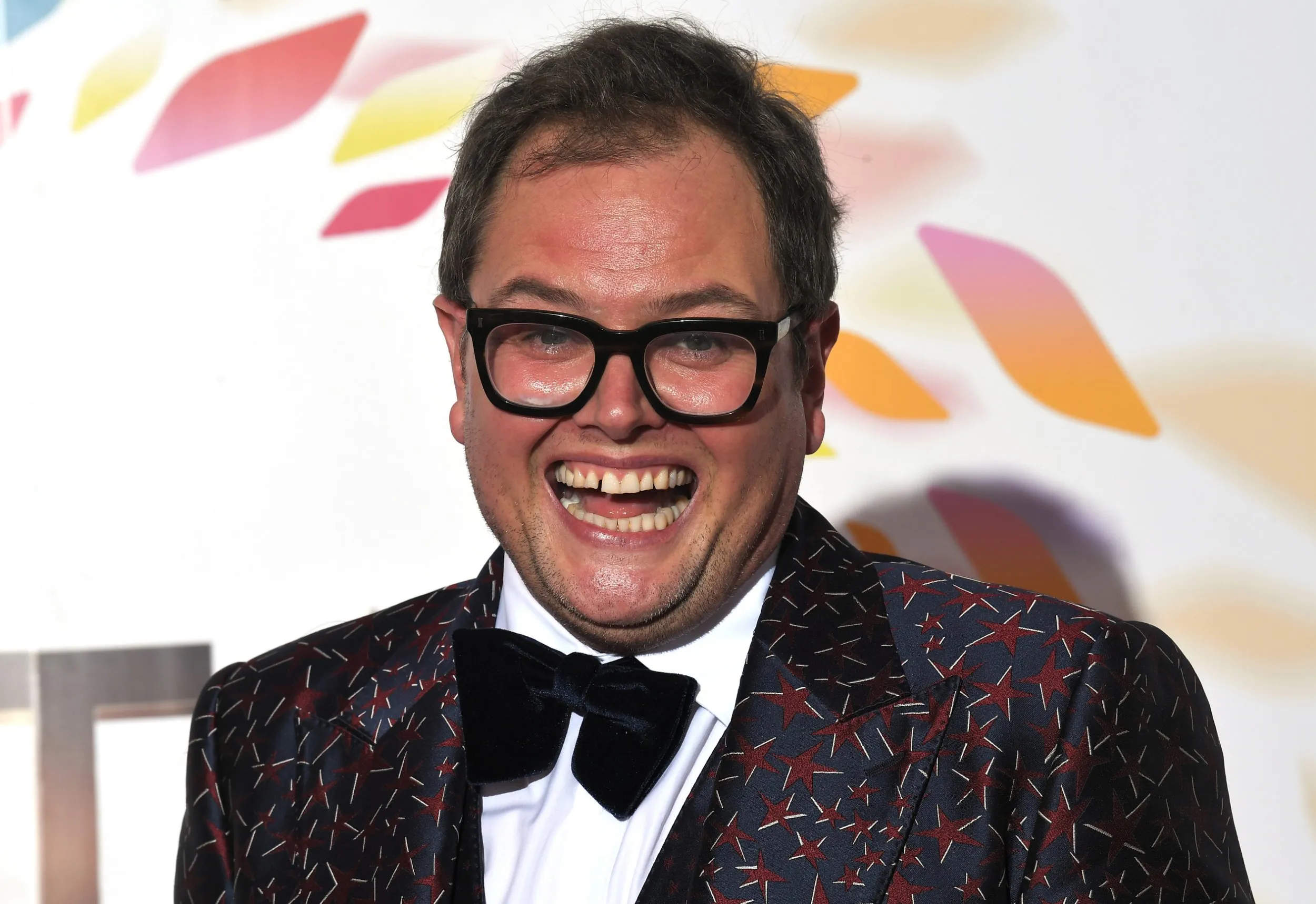 Alan Carr smiling and posing confidently on the red carpet, dressed in a stylish suit with bright lights and cameras in the background.