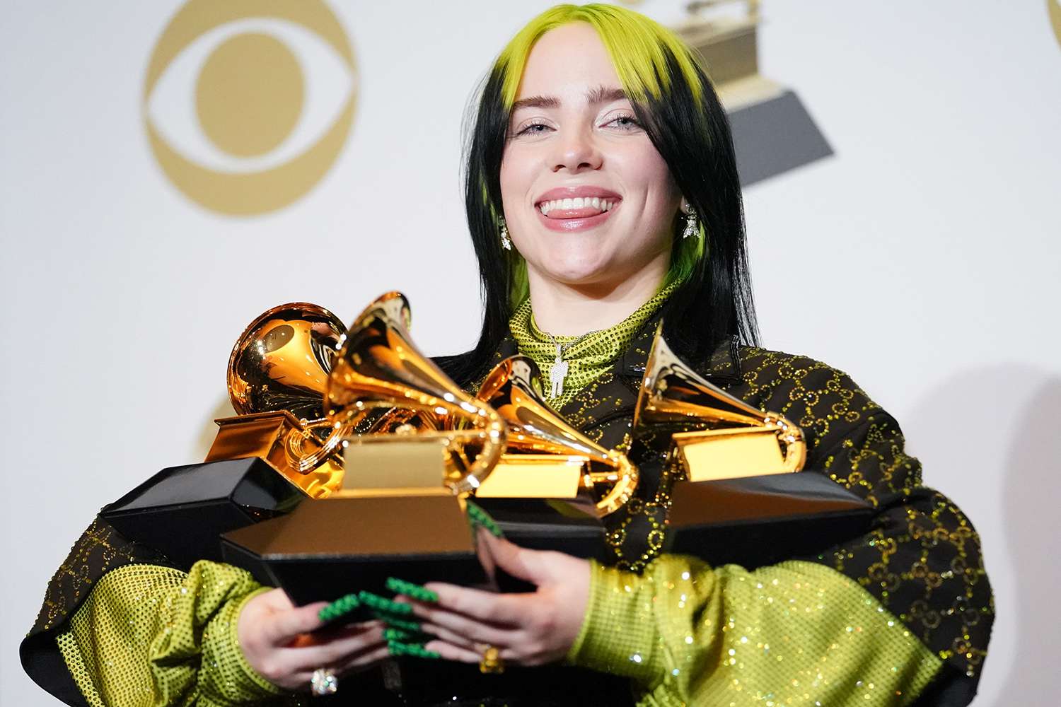 Billie Eilish smiling while holding multiple Grammy Awards after her historic wins, wearing a black and neon-green outfit at the Grammy Awards ceremony.
