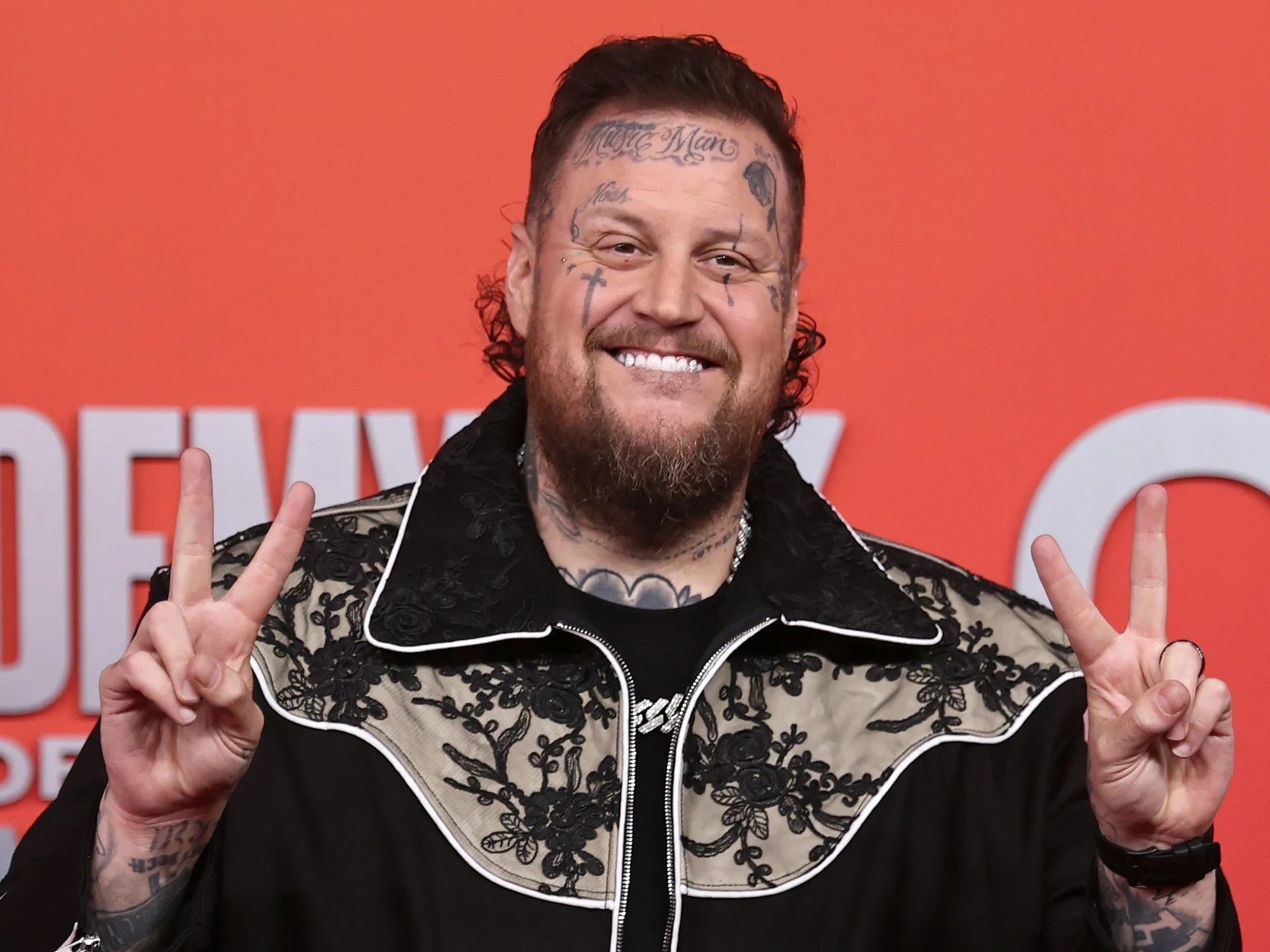 Jelly Roll flashing a peace sign while posing on the red carpet, wearing a black suit and sunglasses.