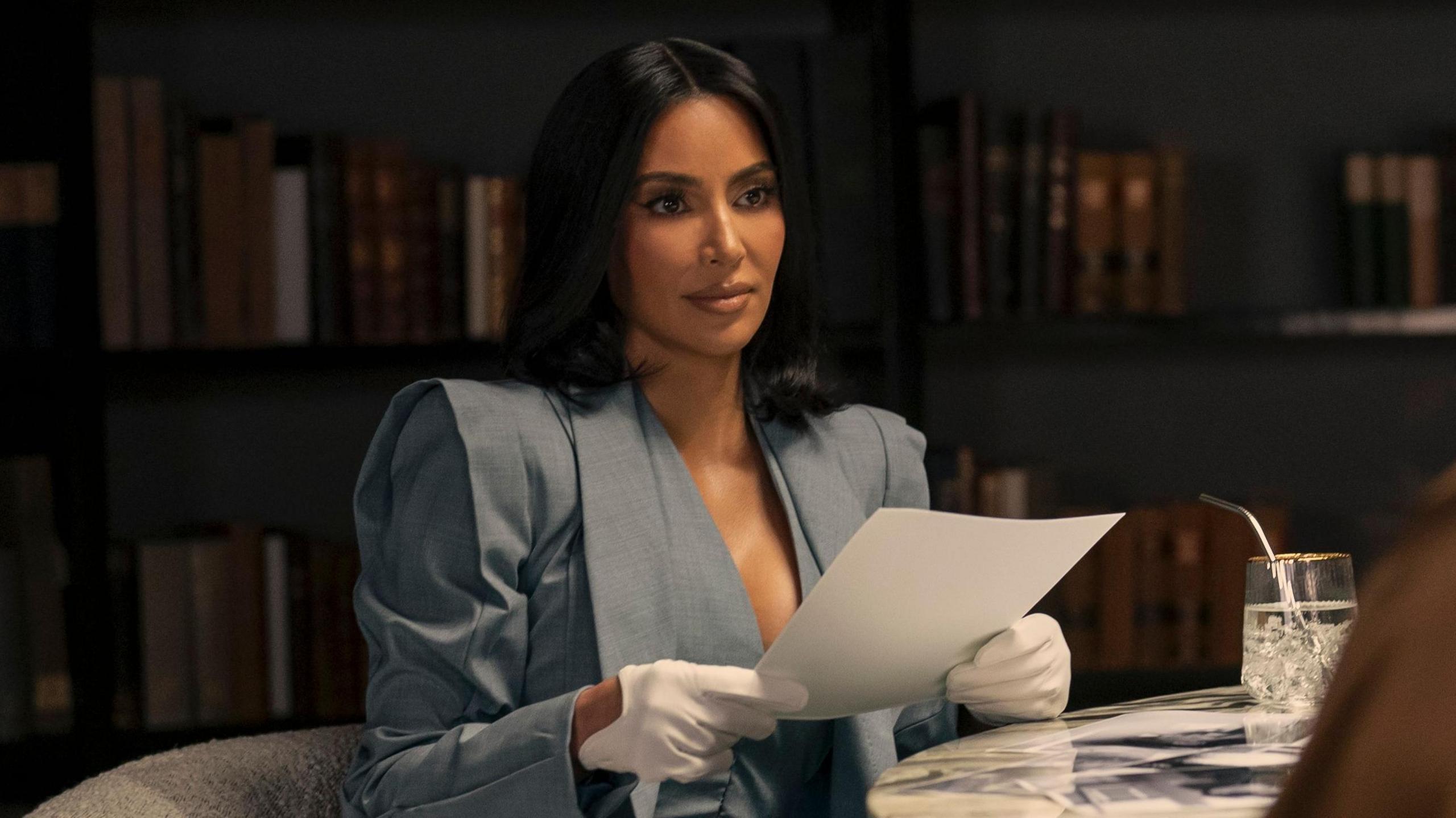 Kim Kardashian sitting at her desk in a grey suit as Allura Grant in Disney+ legal drama All’s Fair, looking focused and professional.