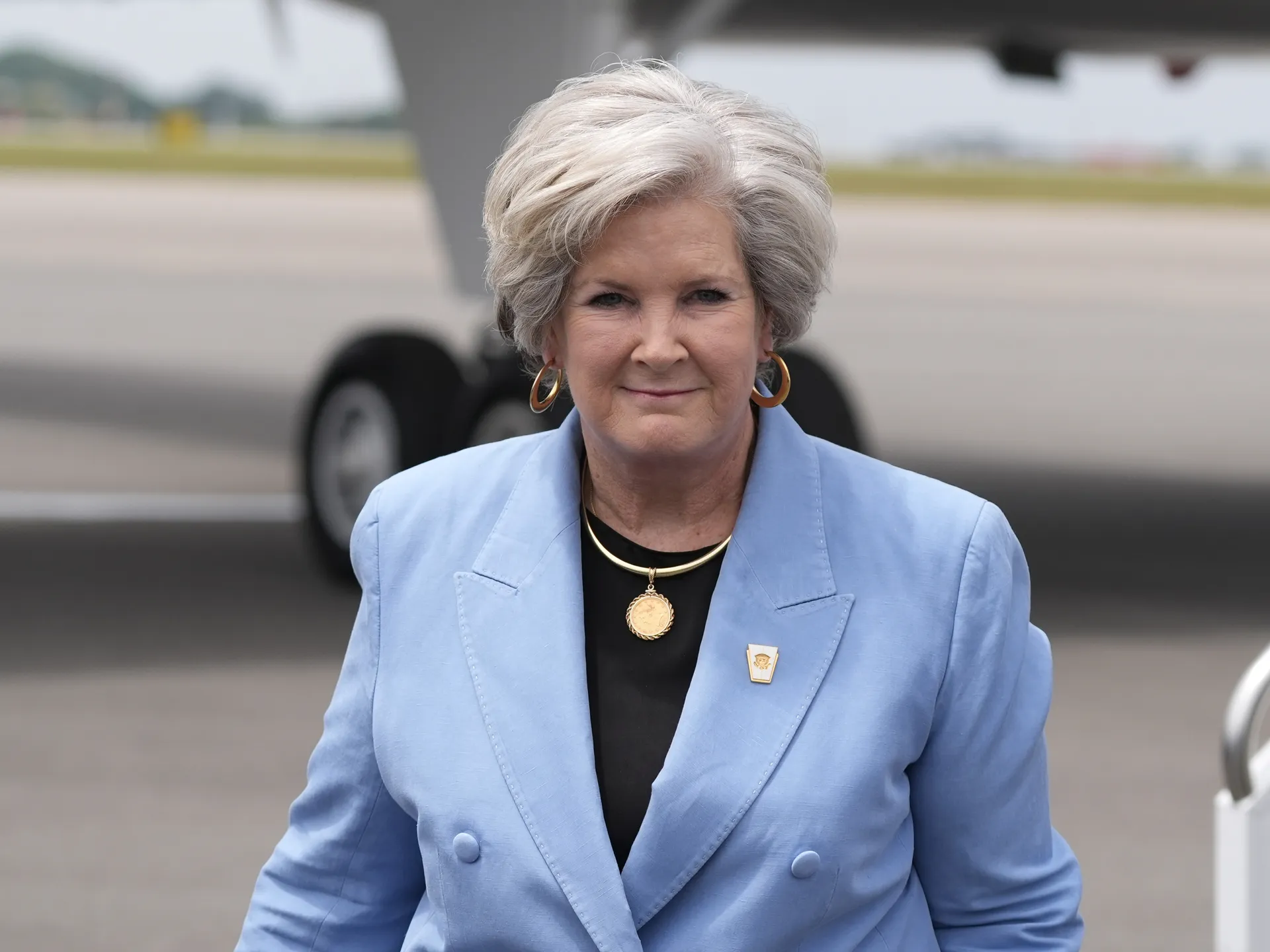 Susie Wiles smiling at the camera as she walks toward Air Force One, wearing a professional outfit.