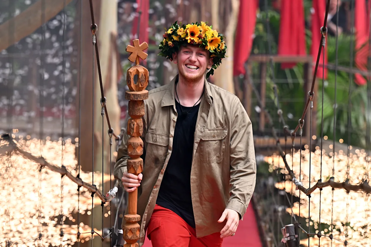 Angry Ginge walks triumphantly across the jungle winner’s bridge after being crowned King of the Jungle 2025, smiling through tears as fireworks light the night sky behind him.