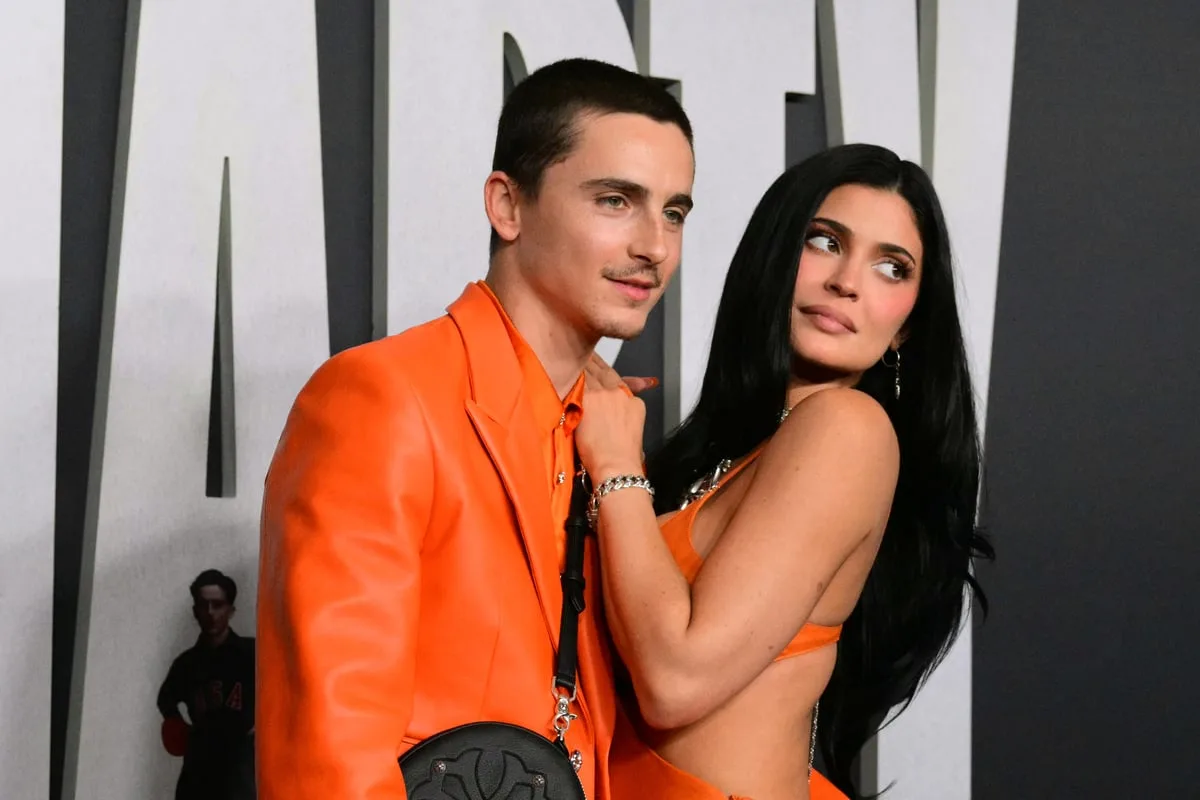 Kylie Jenner and Timothée Chalamet cuddling and smiling together on the red carpet at the Marty Supreme Los Angeles premiere, both wearing coordinated bright orange outfits.