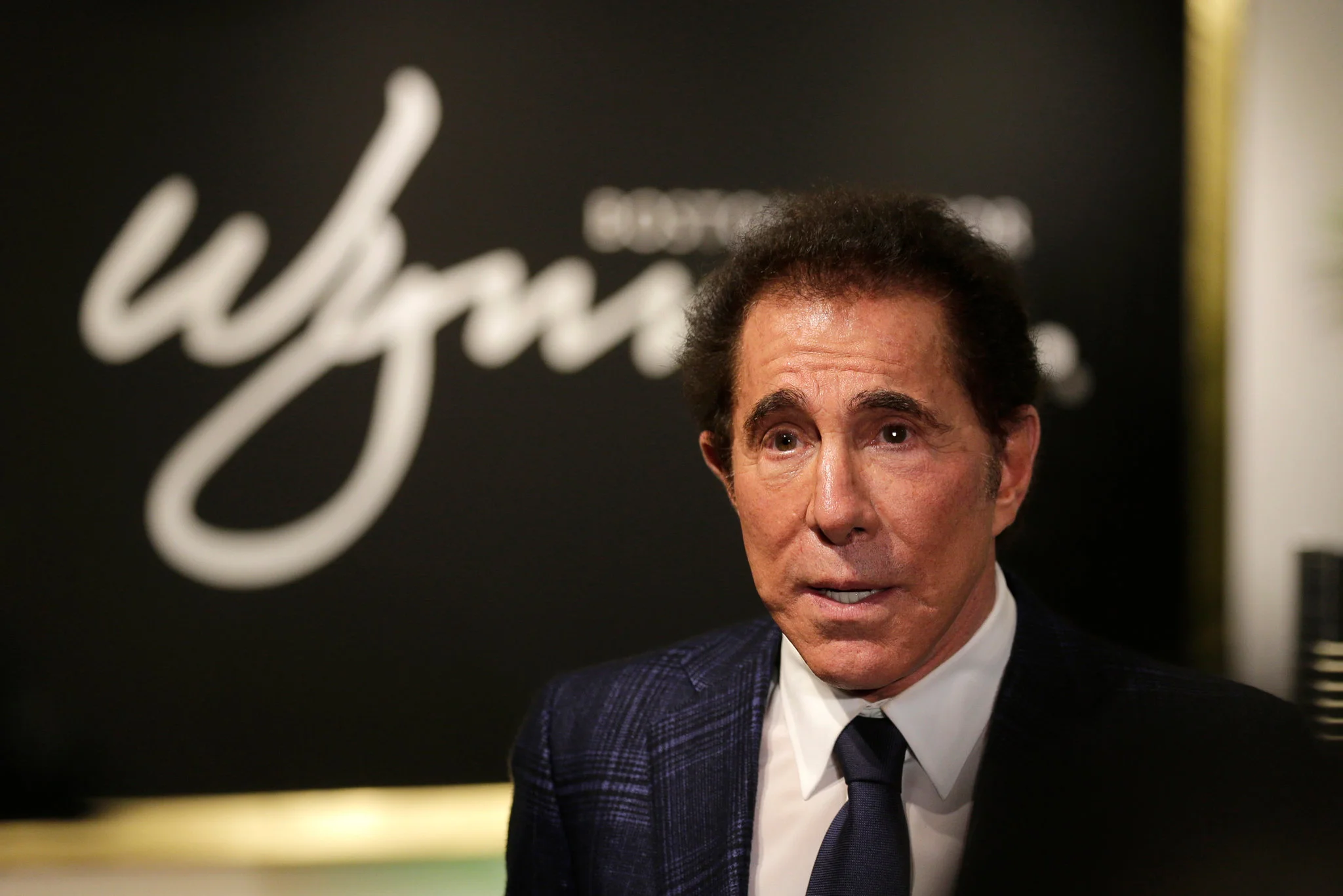 Steve Wynn inside a brightly lit casino, standing near slot machines and tables, surveying the floor in a tailored suit.