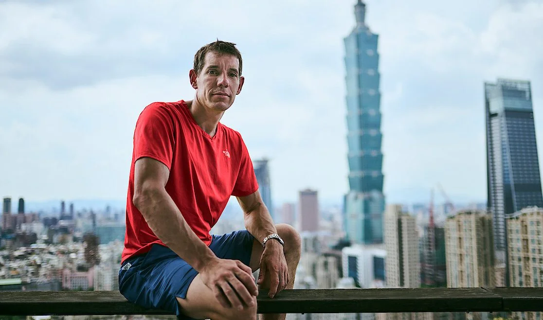 Alex Honnold sits on a ledge overlooking a city skyline, wearing a red shirt with Taipei 101 visible in the background.